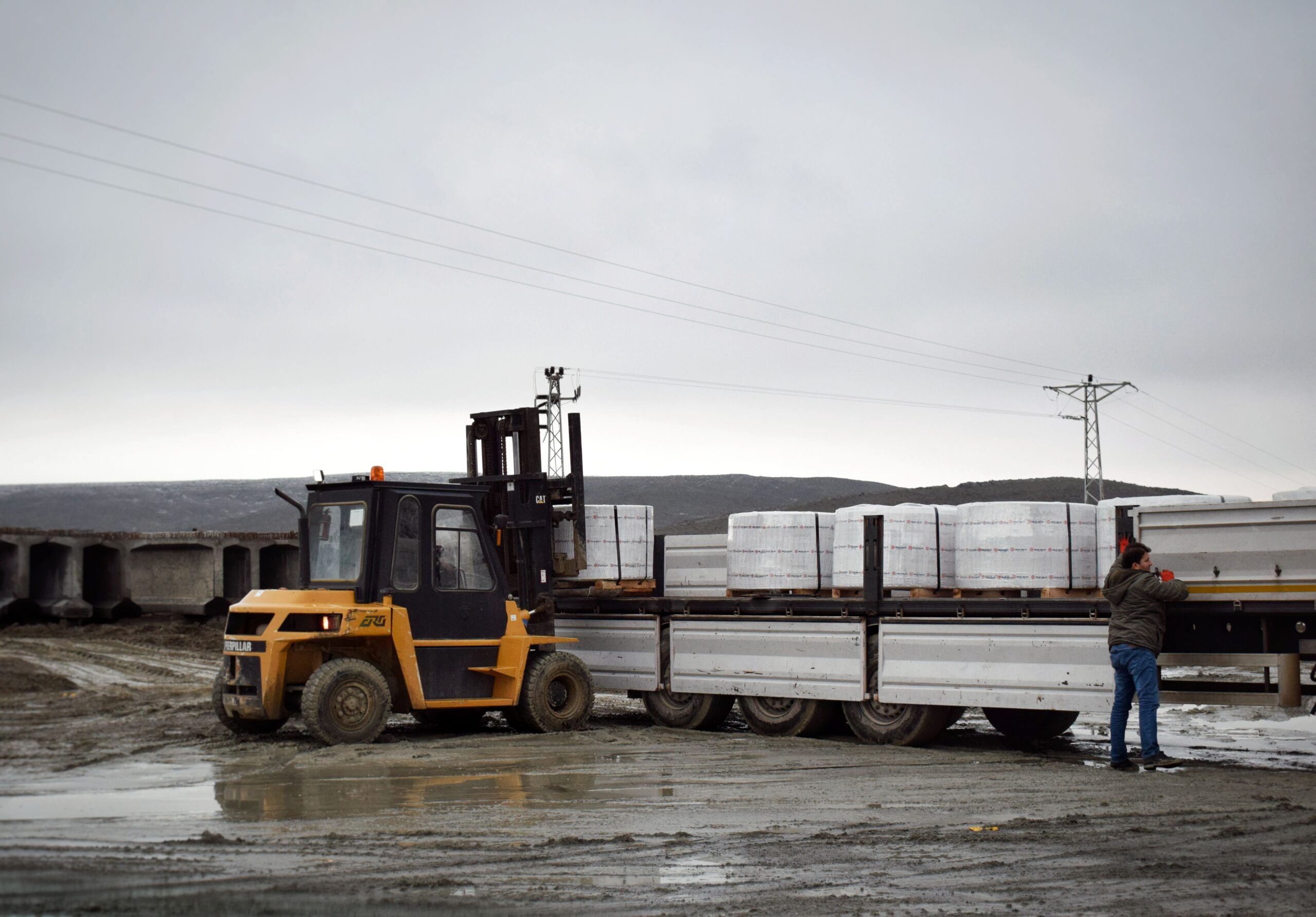 Services A forklift is loading cargo onto a truck in a muddy logistics area with a worker assisting.